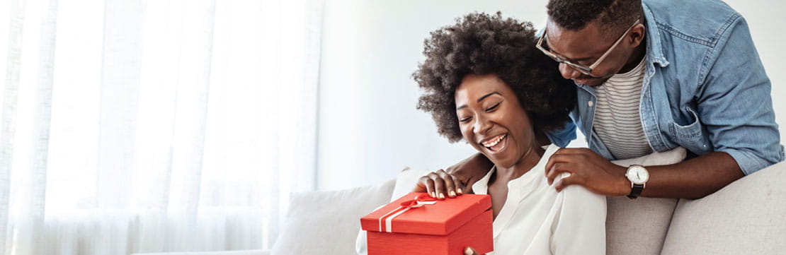 A woman opening a present given by her partner, he can be seen standing behind her.